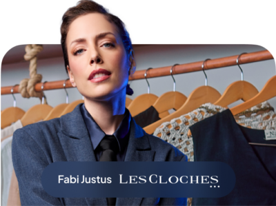 Imagem não clicável: Fabi Justus, dona da marca Les Cloches. Mulher branca com cabelo curto, olhos verdes, blaser azul, camisa azul e gravata preta, ao fundo cabide com roupas de sua marca